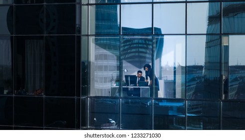 Aerial Shot From Outside Of The Skyscraper: Businessman And Businesswoman Talking Business While Sitting At The Desk By The Office Window. Shot Of The Financial District And Businesspeople Working.