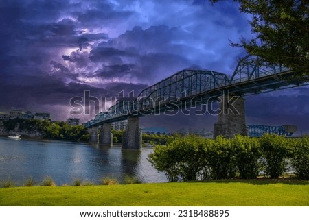 an aerial shot of the Walnut Street Bridge over the rippling blue waters of the Tennessee River surrounded by lush green trees and buildings with powerful storm clouds and lightning at Coolidge park	