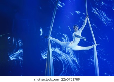 Aerial silk acrobatic performance featuring a strong and flexible woman dressed in a white costume embellished with rhinestones. She is performing tricks in the air, illuminated by blue light. - Powered by Shutterstock