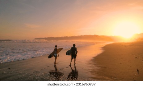 AERIAL: Los surfistas observan las grandes olas que salen de la playa después de las sesiones de surf al atardecer. Magníficas vistas de la orilla de arena naranja brillante a orillas del Océano Pacífico. Surf buddies en una excursión a la playa tropical Venao.