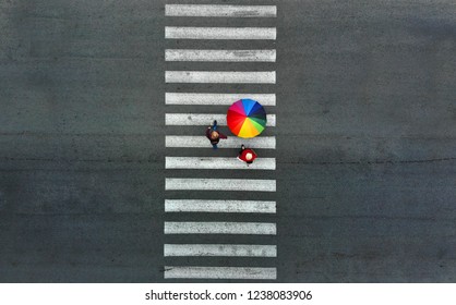 Aerial. Three Pedestrians Walking On A Pedestrian Crossing. One Of Them Holds A Rainbow Umbrella. 