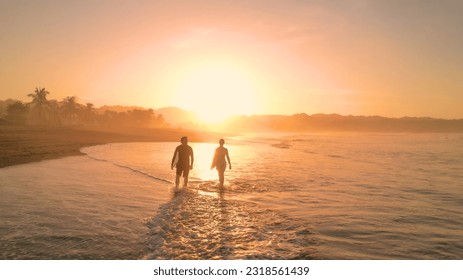 AERIAL: Tidal wave washes on sandy beach and crosses path of two walking surfers. Amazing sun reflection on wet sandy foreshore with surf couple carrying their surfboards in golden sunset light. - Powered by Shutterstock