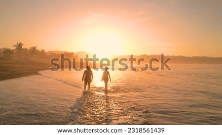 AERIAL: Tidal wave washes on sandy beach and crosses path of two walking surfers. Amazing sun reflection on wet sandy foreshore with surf couple carrying their surfboards in golden sunset light.