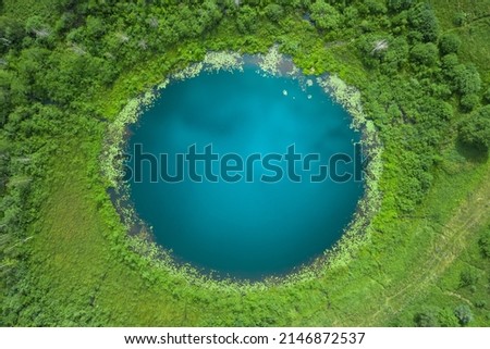Aerial top down amazing lake of round shape. Cloudy sky reflected in clear turquoise water of pond surrounded by trees and plants. Ripple on water surface, windy sunny summer day