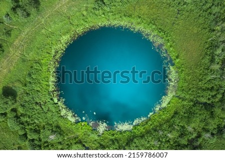 Aerial top down amazing lake of round shape. Cloudy sky reflected in clear turquoise water of pond surrounded by trees and plants. Ripple on water surface, windy sunny summer day.