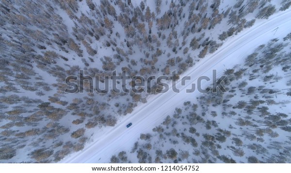 AERIAL TOP DOWN Blue car driving on slippery\
snowy road past lush green frosty spruce trees in amazing dense\
forest. People in auto on road trip across picturesque winter\
landscape in Lapland,\
Finland