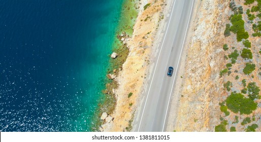 AERIAL, TOP DOWN: Car Cruises Down The Empty Asphalt Road Leading Along The Beautiful Rocky Beach In Lefkas. Flying Above The Coastline Of An Isle In The Mediterranean And Car Driving Down Scenic Road