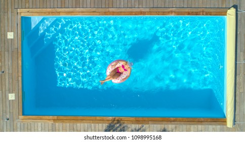 AERIAL, TOP DOWN: Cheerful Young Woman Sunbathing While Relaxing On A Cool Inflatable Doughnut In Her Backyard Pool. Smiling Blonde Haired Girl In Pink Bikini Lying On A Funny Floatie In Amazing Pool.