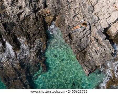AERIAL, TOP DOWN: Female tourist soaks up the Dalmatian sunshine while lying on the scenic wild beach. Relaxed young Caucasian woman on summer vacation on Hvar island sunbathes on the rocky shore.