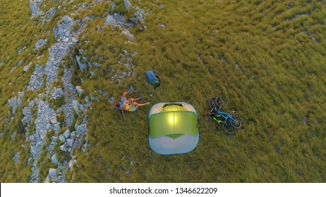 AERIAL TOP DOWN: Flying above the carefree tourist couple observing the evening landscape after a mountain biking journey in Alps. Active man and woman cuddling by their tent in the scenic mountains. - Powered by Shutterstock