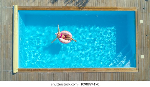 AERIAL, TOP DOWN: Happy Caucasian Woman Paddling Around The Crystal Clear Pool On Her Cool Inflatable Doughnut. Joyful Female Tourist Relaxing On A Funny Floatie During A Day Off At A Luxury Pool.