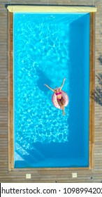 AERIAL, TOP DOWN: Joyful Fit Girl In Pink Swimsuit Chilling On A Doughnut Floatie In The Summer. Gorgeous Caucasian Woman Suntans While Floating Around The Crystal Clear Pool On Inflatable Doughnut.