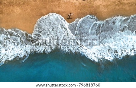 AERIAL, TOP DOWN: Unrecognizable surfer walking along the exotic sandy beach on sunny summer day. Lonely man holding surfboard looking at splashing ocean waves. Splashing ocean waves reach sandy beach