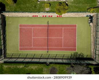 Aerial top down view of an empty red tennis court with white lines and a net, surrounded by green artificial grass and a fence on a sunny day.