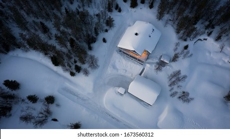 Aerial Top Down View Of A House Covered In Snow In The Woods With Cozy Lights In The Building