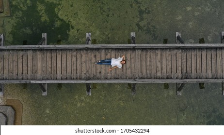 Aerial Top Down View Of Young Woman Lying On The Wooden Bridge Over Mountain River, Pond. Hallstatt, Austria.