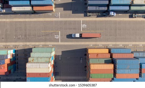 AERIAL, TOP DOWN: White truck transports a red freight container across the busy port of Los Angeles. Flying above a freight truck hauling a heavy shipment past large stacks of other containers.
