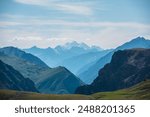 Aerial top view above green pass to rocky deep gorge with sharp sheer crags silhouettes against huge snowy mountain under clouds in blue sky. Dramatic layered scenery with giant snow castle far away.