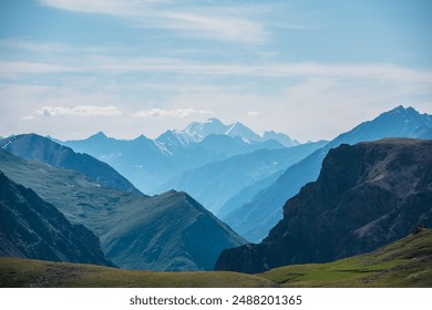 Aerial top view above green pass to rocky deep gorge with sharp sheer crags silhouettes against huge snowy mountain under clouds in blue sky. Dramatic layered scenery with giant snow castle far away. - Powered by Shutterstock