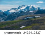 Aerial top view to big snow castle in bright sun under clouds in blue sky. Scenic landscape with sunlit green hills with snowfields against large snow-white rocky mountain peak far away in sunny day.