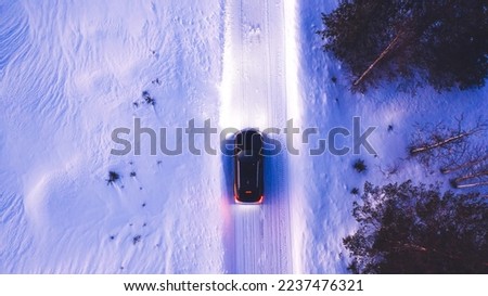 Aerial top view of black car driving on winter road in rural area while headlights illuminate in the dark, bird's eye view of suv vehicle crossing snowy cold lands with insurance. Lighting the way