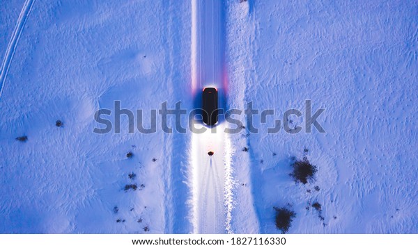 Aerial top view of car on rural area road while\
headlights are on in winter darkness, bird\'s eye view of suv\
vehicle in snowy north lands. Person standing front automobile\
which lighting the way