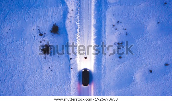 Aerial top view of car on rural area road while
headlights are on in winter darkness, bird's eye view of suv
vehicle in snowy north lands. Person standing front automobile
which lighting the way