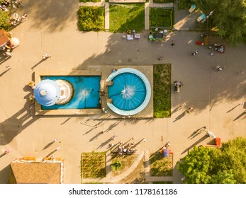 Aerial Top View Of Public City Summer Park With Crowd Of People Walking By The Fountains