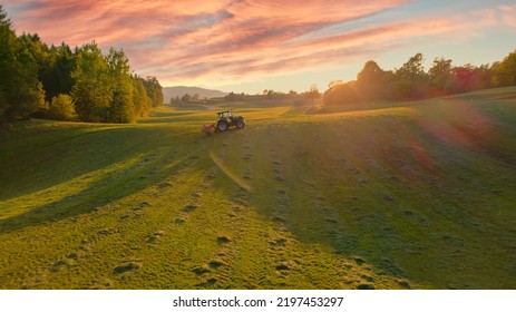 AERIAL: Tractor turning over mowed hay on a sunny autumn morning in golden light. Farmer using hay tedder to aerate grass and speed up drying. Preparing fodder for cows as mandatory farm task in fall. - Shutterstock ID 2197453297