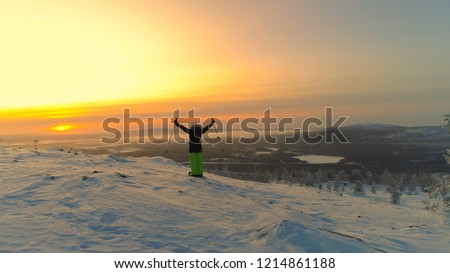 AERIAL: Unrecognizable female skier looks over the spectacular Lapland at sunset. Young woman celebrates reaching the summit on her skis on a sunny winter evening. Picturesque wintry nature in Finland