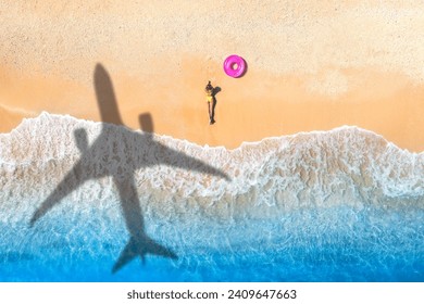 Aerial view of airplane shadow, lying beautiful young woman, pink swim ring, tropical sandy beach, sea with waves at sunset. Summer vacation in island. Top view of slim girl, azure water, plane shadow