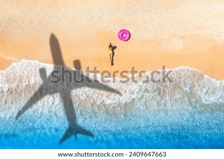 Aerial view of airplane shadow, lying beautiful young woman, pink swim ring, tropical sandy beach, sea with waves at sunset. Summer vacation in island. Top view of slim girl, azure water, plane shadow