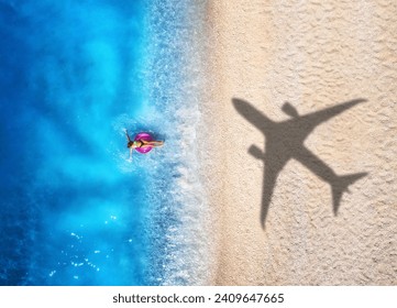 Aerial view of airplane shadow, swimming beautiful young woman with pink swim ring, tropical sandy beach, sea with waves at summer sunny day. Vacation. Top view of slim girl, azure water, plane shadow