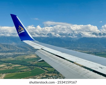 An aerial view from an airplane window, showing the wing of a plane in flight. A landscape unfolds with small towns, fields, and greenery. Snow-capped mountains, partially covered by clouds. - Powered by Shutterstock