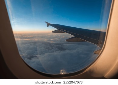 An aerial view from an airplane window showing a wing over a blanket of clouds, with a gradient sunset or sunrise casting warm orange to deep blue hues across the sky - Powered by Shutterstock