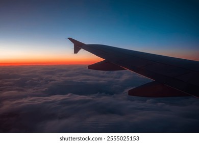 An aerial view from an airplane window showing a wing over a blanket of clouds, with a gradient sunset or sunrise casting warm orange to deep blue hues across the sky - Powered by Shutterstock