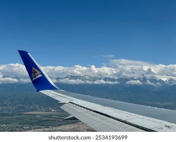 An aerial view from an airplane window, wing of a plane in flight. A picturesque landscape with small towns, fields, and greenery. In the distance, snow-capped mountains, partially covered by clouds. - Powered by Shutterstock