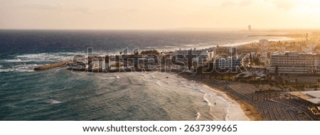 Aerial view of Ayia Napa, Cyprus, at sunset with sandy beaches, turquoise waters, a small harbor with boats, and luxury hotels in the background.