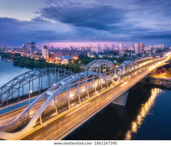 Aerial view of beautiful bridge at night in Kiev,\
Ukraine. Landscape with bridge, river, city illumination, blue sky\
with clouds at sunset. Cityscape with road, cars, buildings, city\
lights. Top view