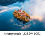 Aerial view of beautiful church and buildings on the island on the famous Bled Lake, Slovenia in autumn morning. Top drone view of chapel, houses, orange trees, blue water in fall. Landscape. Travel