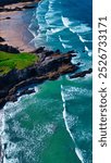 Aerial view of a beautiful coastline featuring sandy beaches, rocky cliffs, and turquoise waves crashing onto the shore. At Porth Beach, Cornwall, UK.