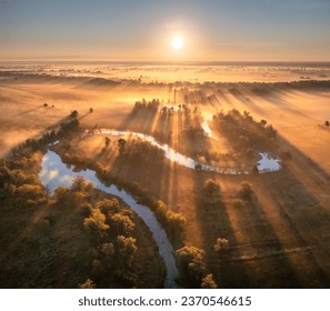 Aerial view of beautiful curving river in low clouds at sunrise in autumn in Ukraine. Turns of river, meadows and fields, grass, orange trees, golden sun rays at dawn in fall. Top view of river coast