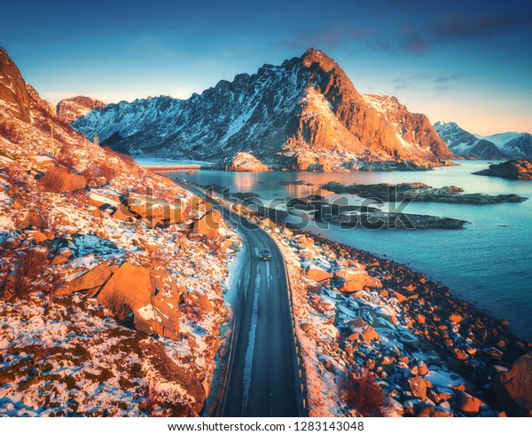 Aerial view of beautiful mountain road near the\
sea, mountains, purple sky at sunset in Lofoten islands, Norway in\
winter. Top view of road, car, high snowy rocks with stones,\
coastline, blue water