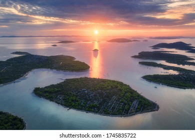 Aerial view of beautiful small islands in Adriatic sea at sunset in summer in Croatia. Top view of blue water, green trees on the mountains, colorful sky with clouds and orange sun. Tropical landscape