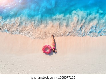 Aerial view of the beautiful young lying woman with pink donut swim ring on the white sandy beach near sea with waves at sunset. Summer holiday. Top view of slim girl, clear azure water. Indian Ocean