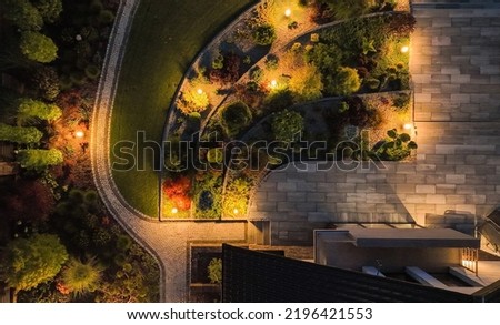 Aerial View of Beautifully Designed and Landscaped Garden on the Backyard of Residential House. Garden Path and Terrace Surrounded by Variety of Plants Enlightened by Solar Lamps. Evening Time.