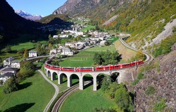 Aerial View Of A Bernina Express Train Crossing The Famous Brusio Spiral Viaduct Of Rhaetian Railway Over A Green Grassy Meadow With Fall Colors On The Rocky Mountainside, In Graubunden, Switzerland