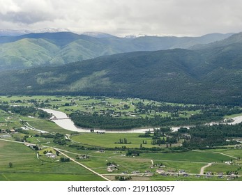 Aerial View Of The Bitterroot River Flowing Through Lush, Green Fields, Next To Homes, Hills And Mountains In Missoula Area, Montana, USA. Rain Clouds Overhead