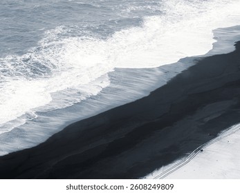 Aerial view of Dyrhólaey black beach in Iceland, where waves meet volcanic sand and fresh snow. Abstract winter coastal landscape with bold contrast and textures