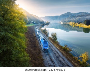 Aerial view of blue modern high speed train moving near river in alpine mountains in fog at sunrise in autumn. Top view of train, railroad, lake, reflection, trees in fall. Railway station in Slovenia